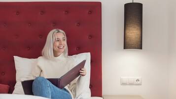 Una persona sentada en una cama con cabecera roja, sosteniendo un libro. Hay una mesita de noche con una lámpara y un enchufe en la pared.