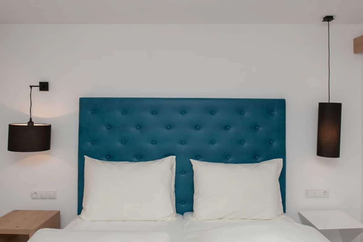 Un dormitorio con cabecera tapizada azul, ropa de cama blanca y dos mesitas de noche con lámparas.