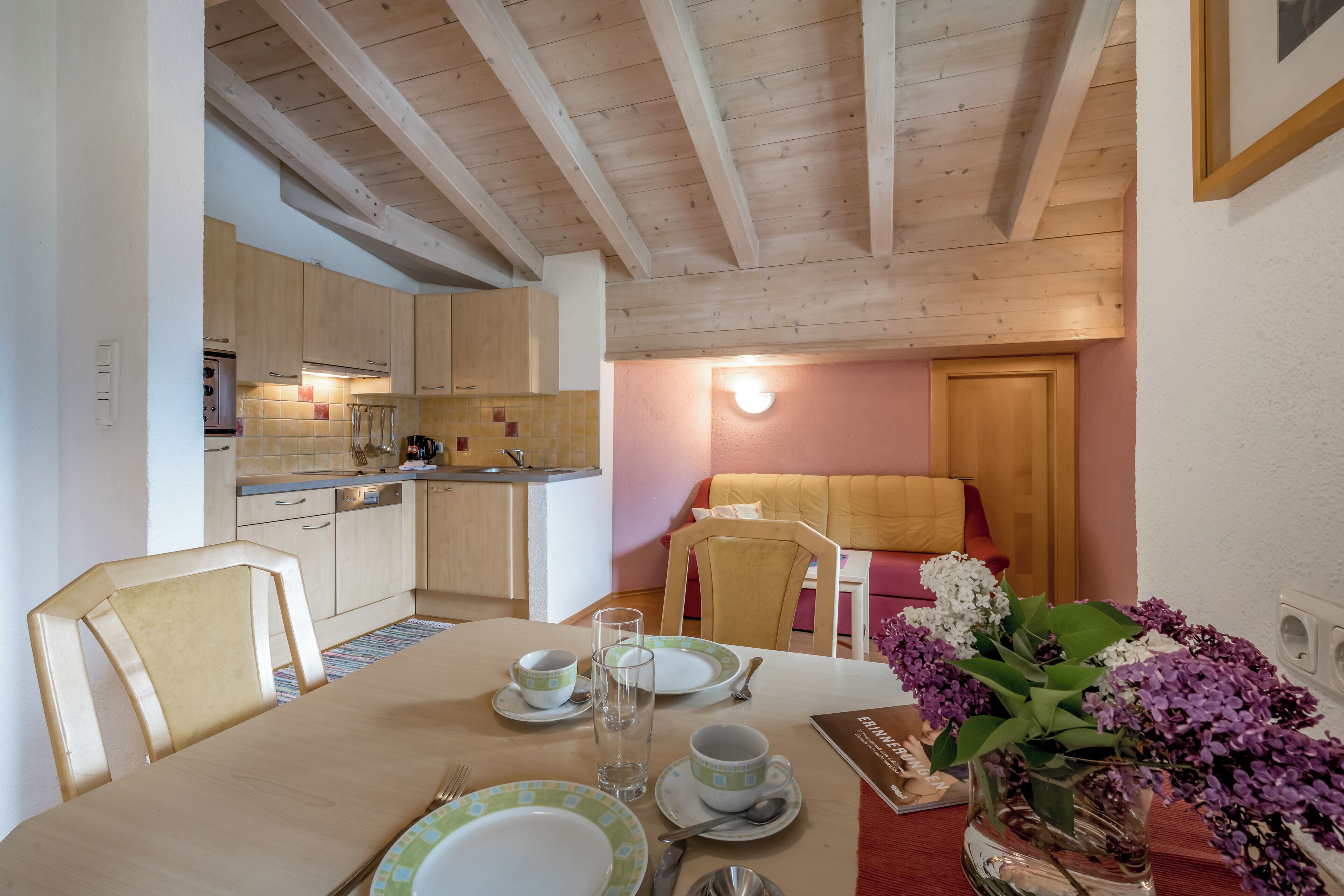 Un comedor con mesa de madera dispuesta para dos personas, una cocina al fondo y un rincón de estar acogedor con sofá y lámpara.