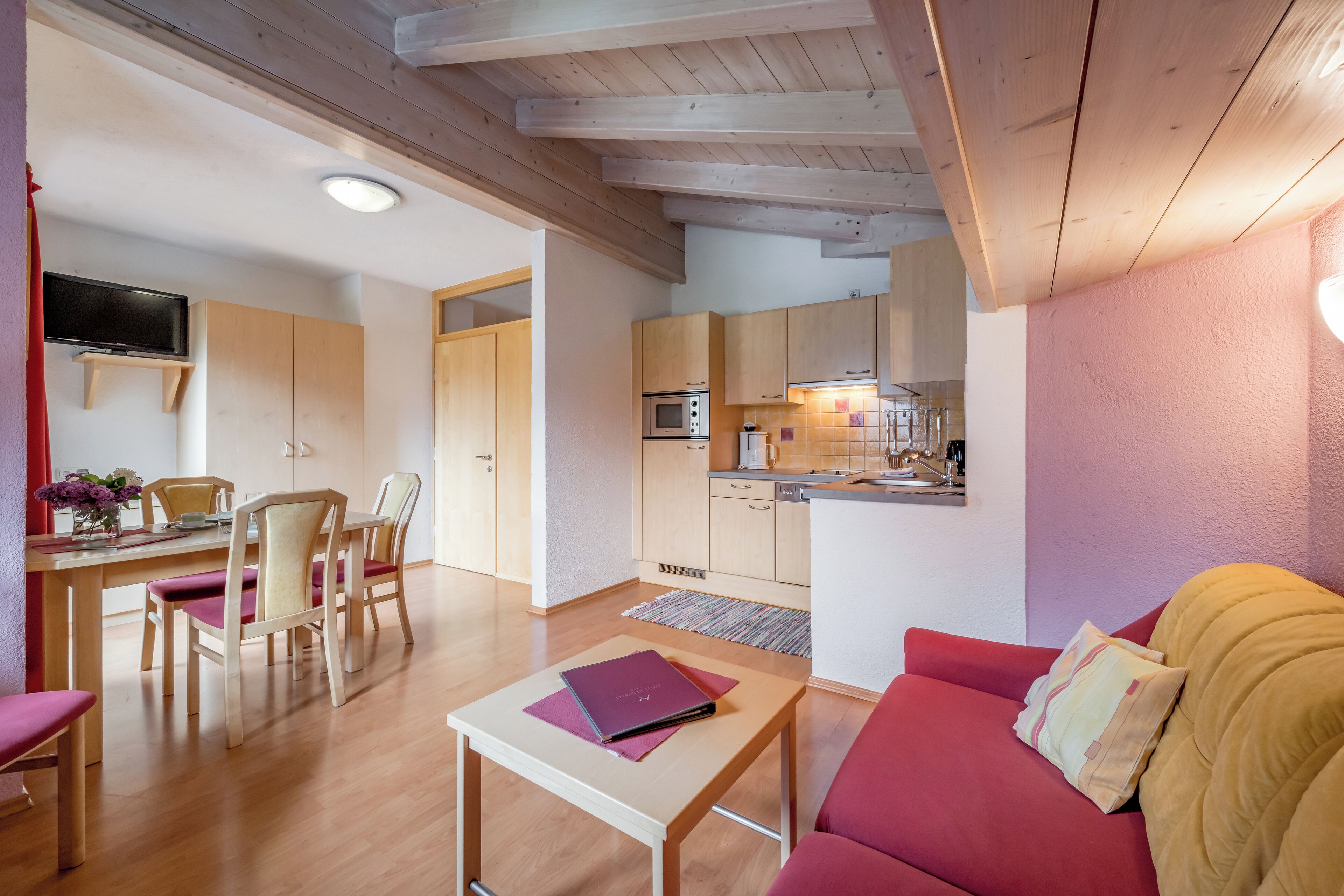 A modern living room with a red sofa, a wooden dining table with chairs, a small table with a book, and a kitchen area with wooden cabinets and a microwave.