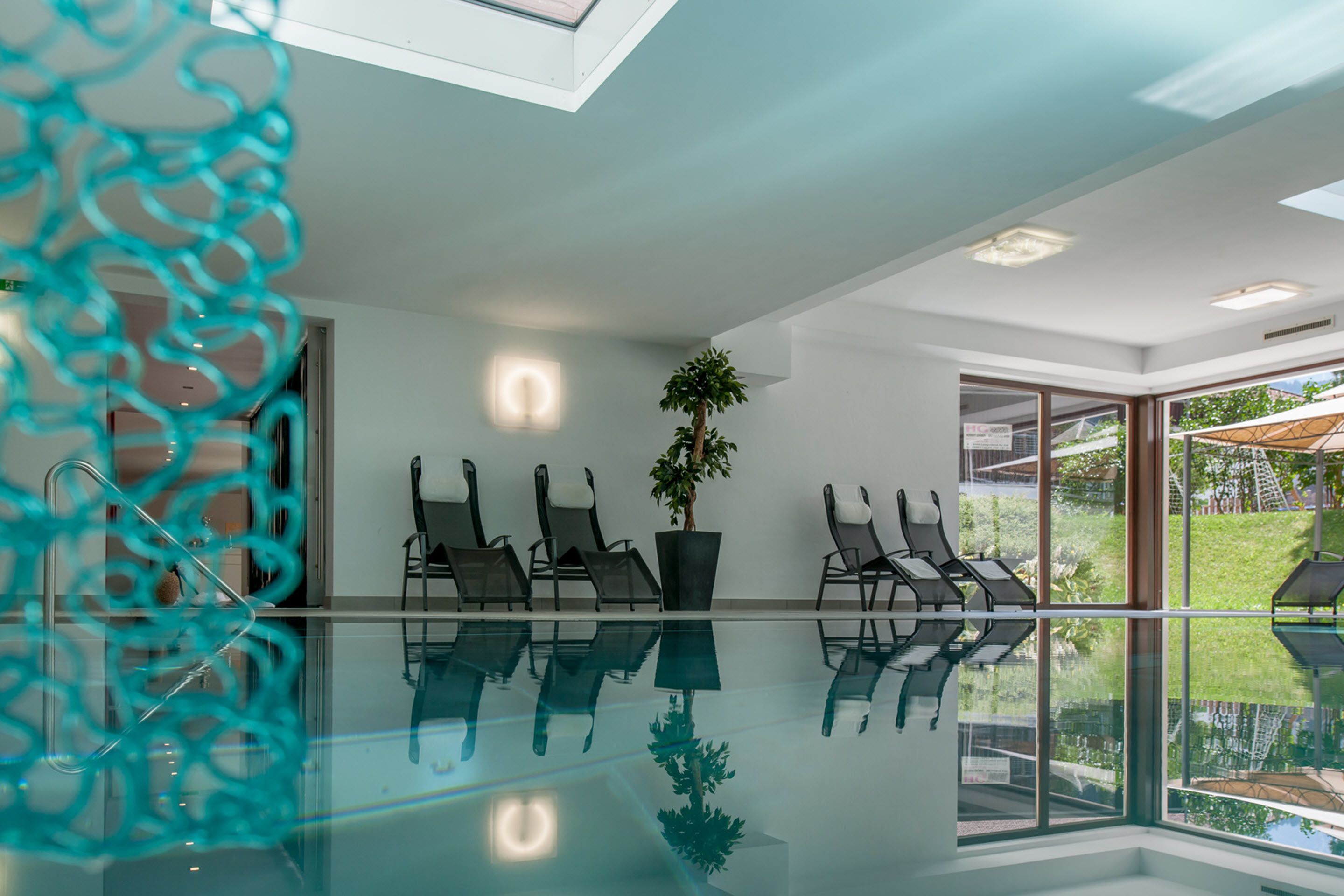 A modern indoor pool area with clear glass walls, a potted plant, and several lounge chairs.