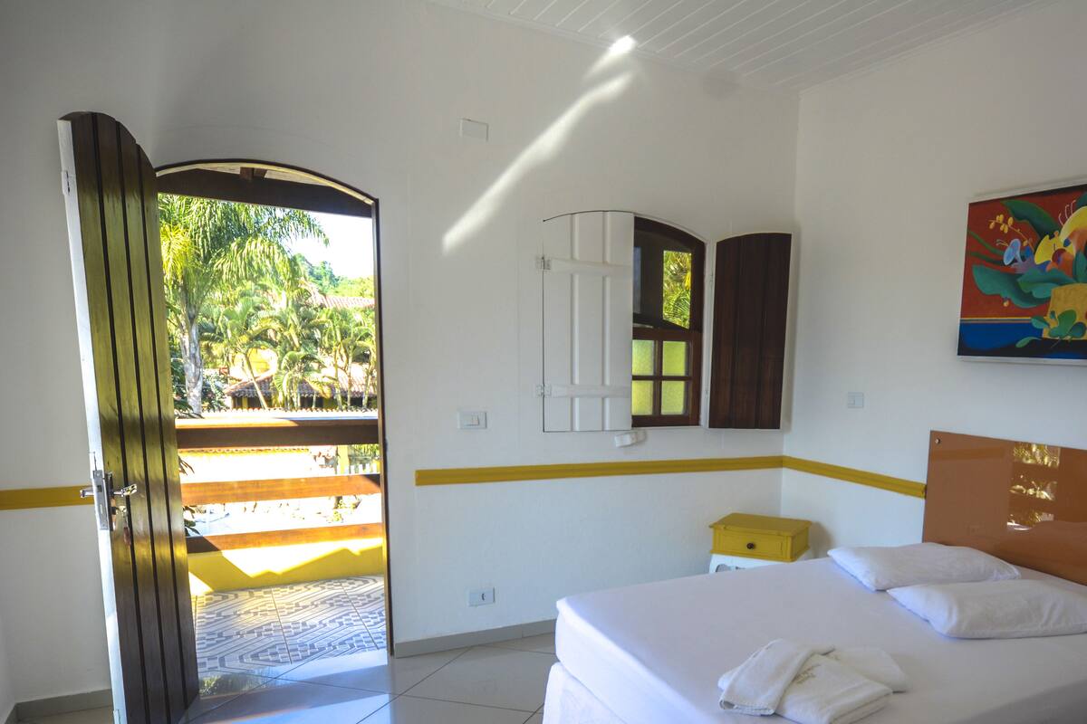 Quarto de hotel com cama, uma pequena mesa de cabeceira, espelho e porta para área externa.