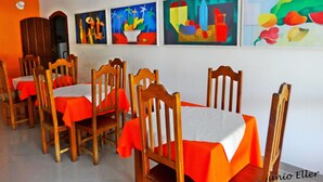 A dining area with wooden chairs and tables, each covered with a white tablecloth and red table runner. There are four colorful paintings on the wall. - Pousada Praia do Jabaquara (Paraty)