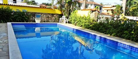 Una piscina rectangular con baldosas azules y un borde de piedra, rodeada de exuberante vegetación y un muro de piedra.