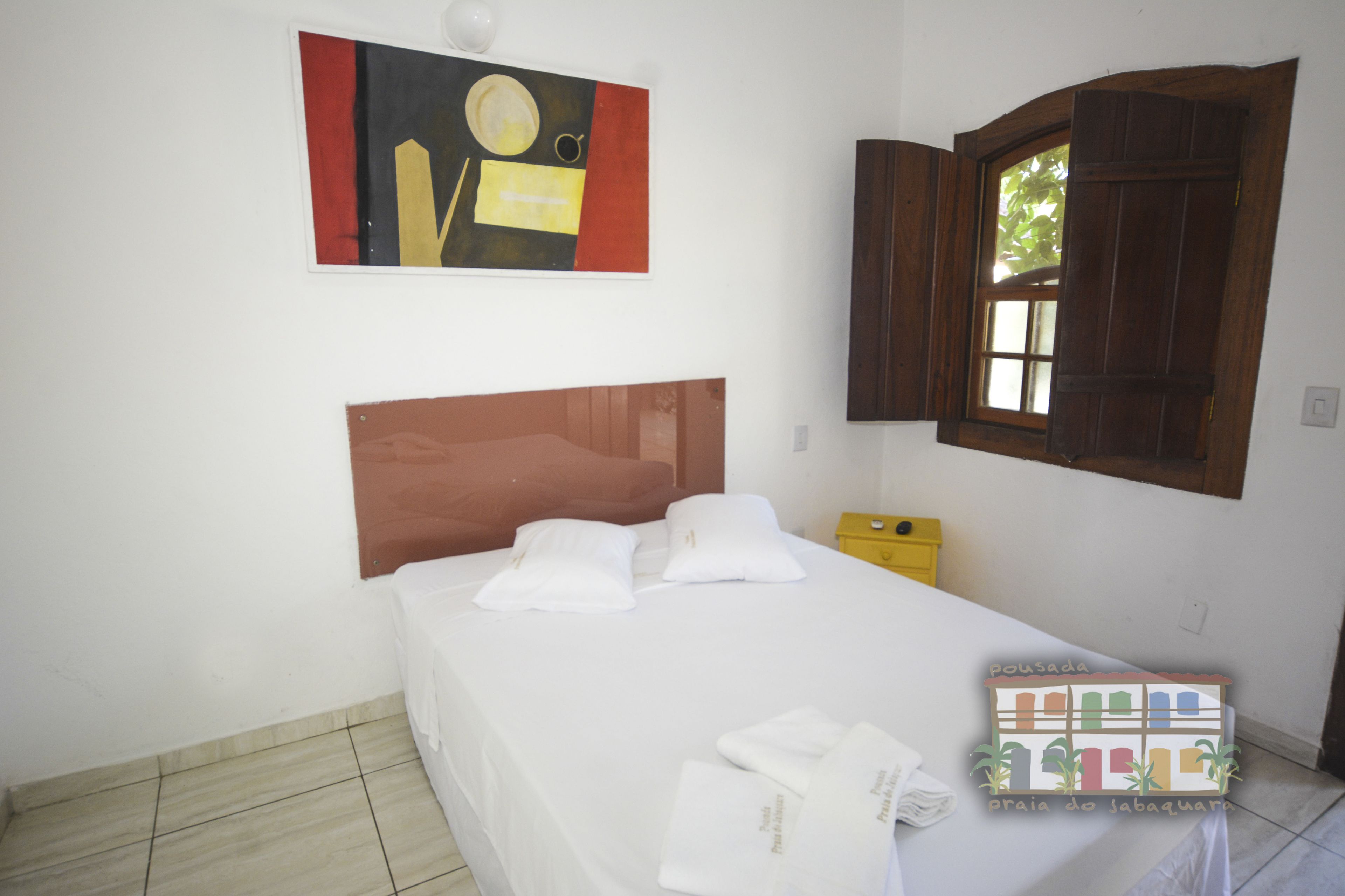 Une chambre d’hôtel avec un lit, une fenêtre avec des volets en bois, un tableau au mur et une boîte jaune sur la table de chevet.