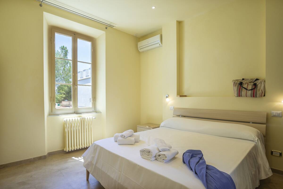 Una habitación de hotel con una cama grande, una ventana, aire acondicionado, una mesita de noche y un estante en la pared.