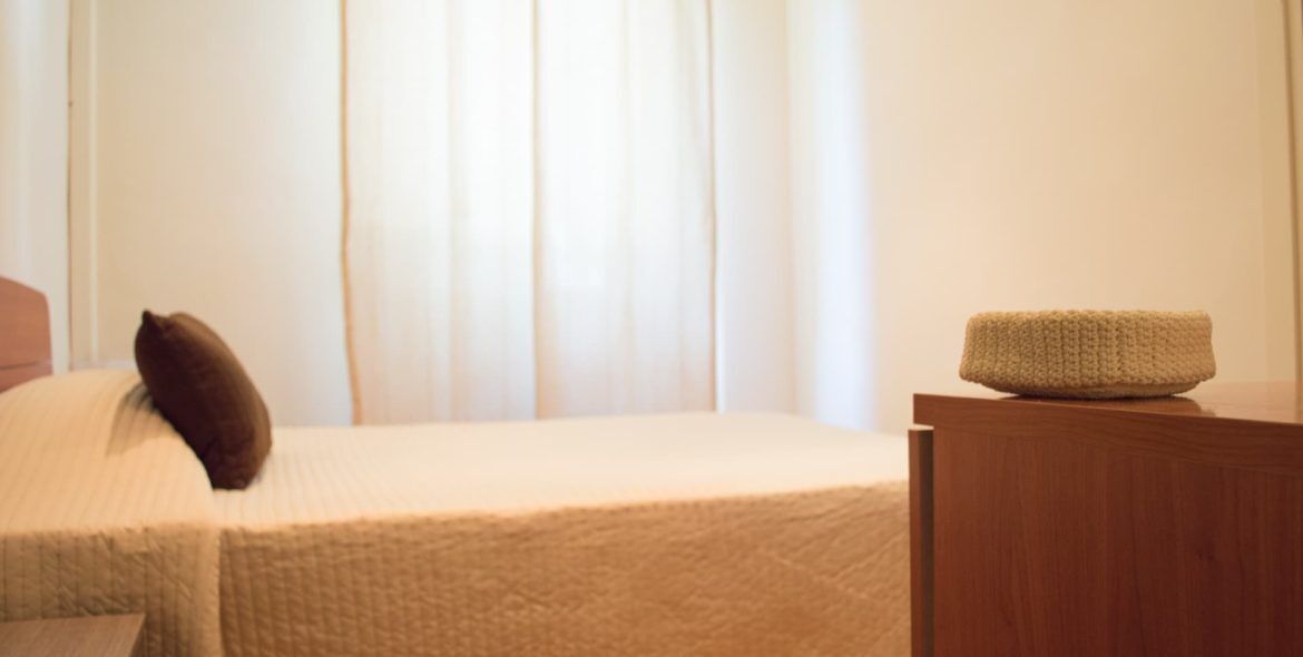 Un letto rifatto con cura, un cuscino, un comodino in legno e un cesto di vimini sopra.