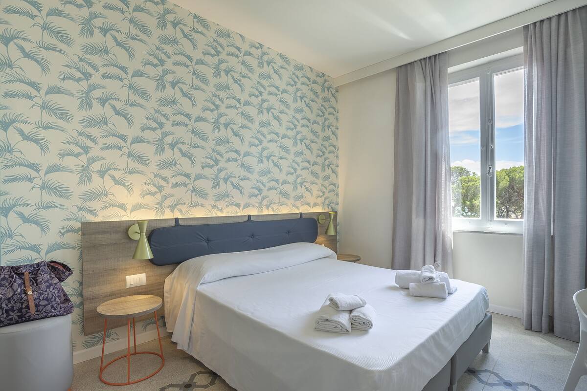 Una cama bien hecha con sábanas y toallas blancas, una mesita de noche con lámpara, y vistas al exterior a través de una ventana con cortinas.