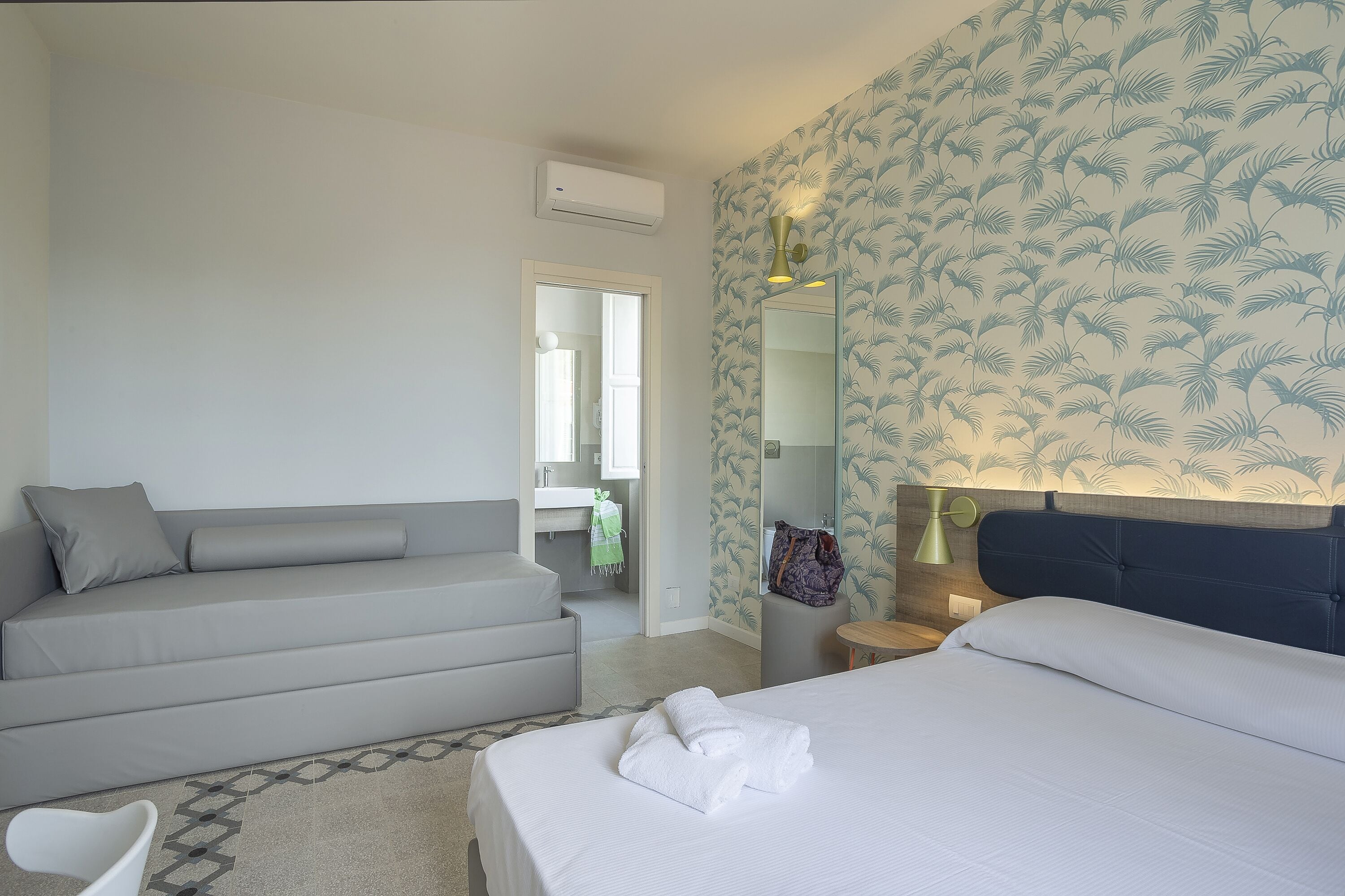 Et moderne hotelværelse med en grå sofa, seng med hvide sengetøj, et lille bord og et spejl.