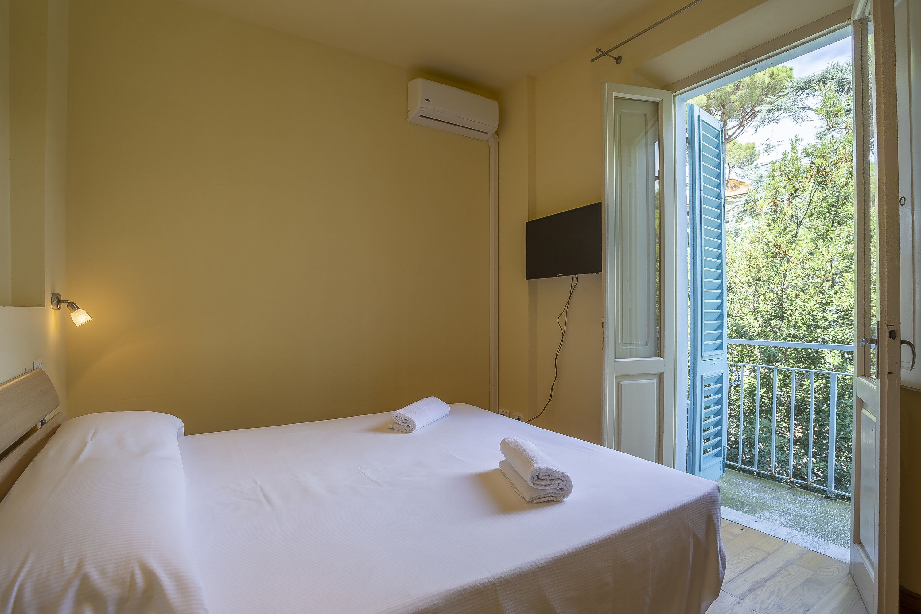 Une chambre avec un lit, deux serviettes, un appareil de climatisation, une télévision et un balcon doté d’une rambarde.
