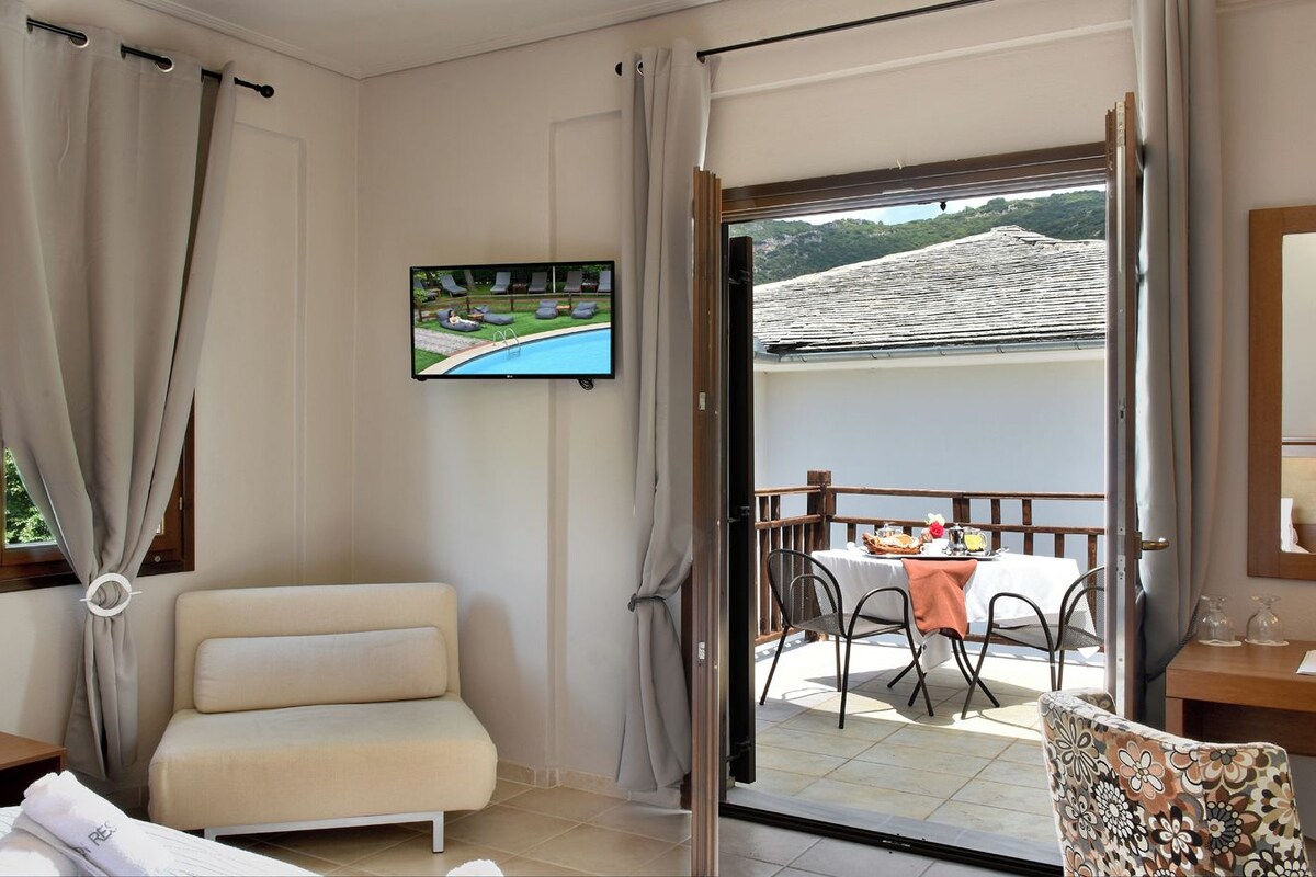A hotel room with a sofa, a dining table with chairs, a TV, and a balcony with a view of a pool and hills.