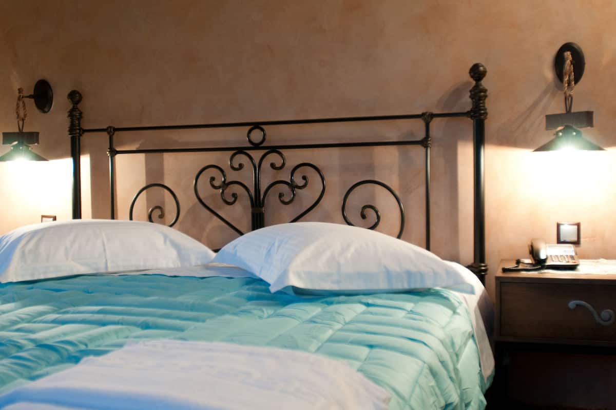 A bed with a teal comforter and white pillows, a black metal headboard, and two bedside lamps on a wooden nightstand.