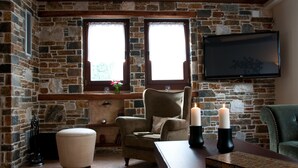 A cozy room with a stone fireplace, a comfortable armchair, a wooden table with candles, and a television mounted on the wall. - Belina Hotel (Volos)
