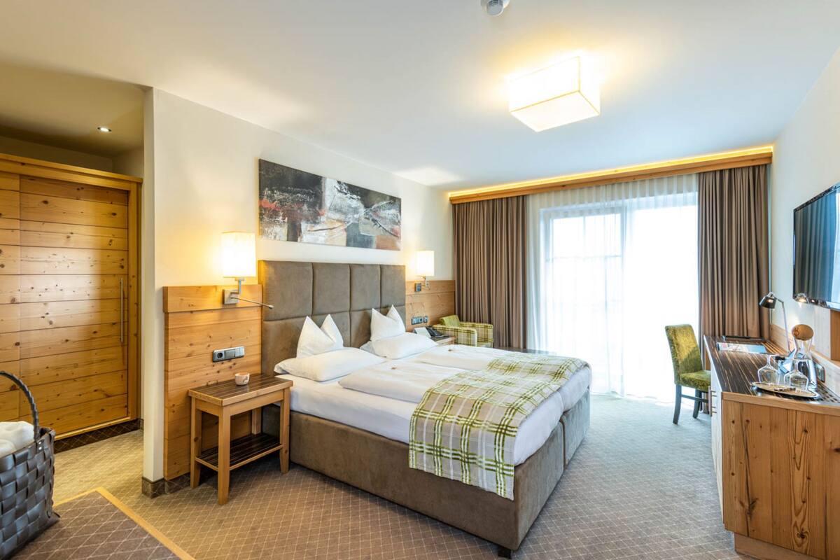 Una habitación de hotel con una cama grande, cabecera de madera, un escritorio con silla, televisión y una ventana con cortinas.