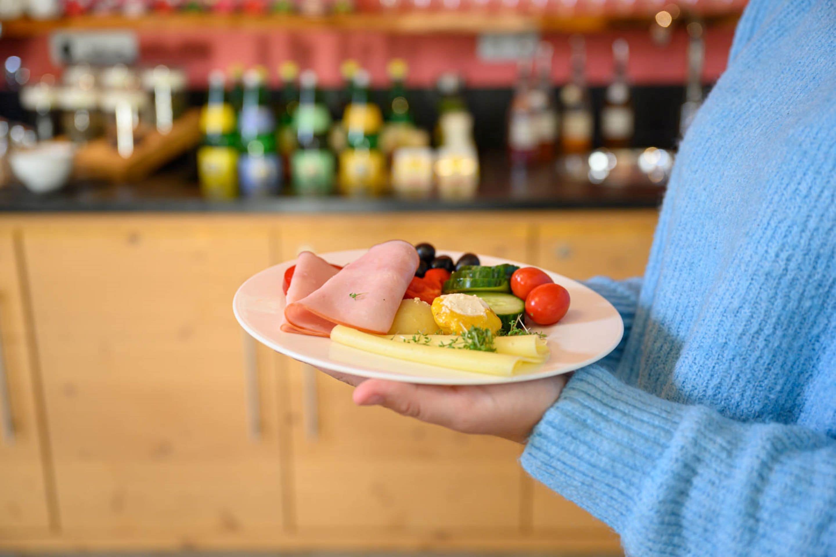 A person holding a plate with various foods, including ham, vegetables, and cheese, in a kitchen setting with wooden cabinets and a bar in the background.
