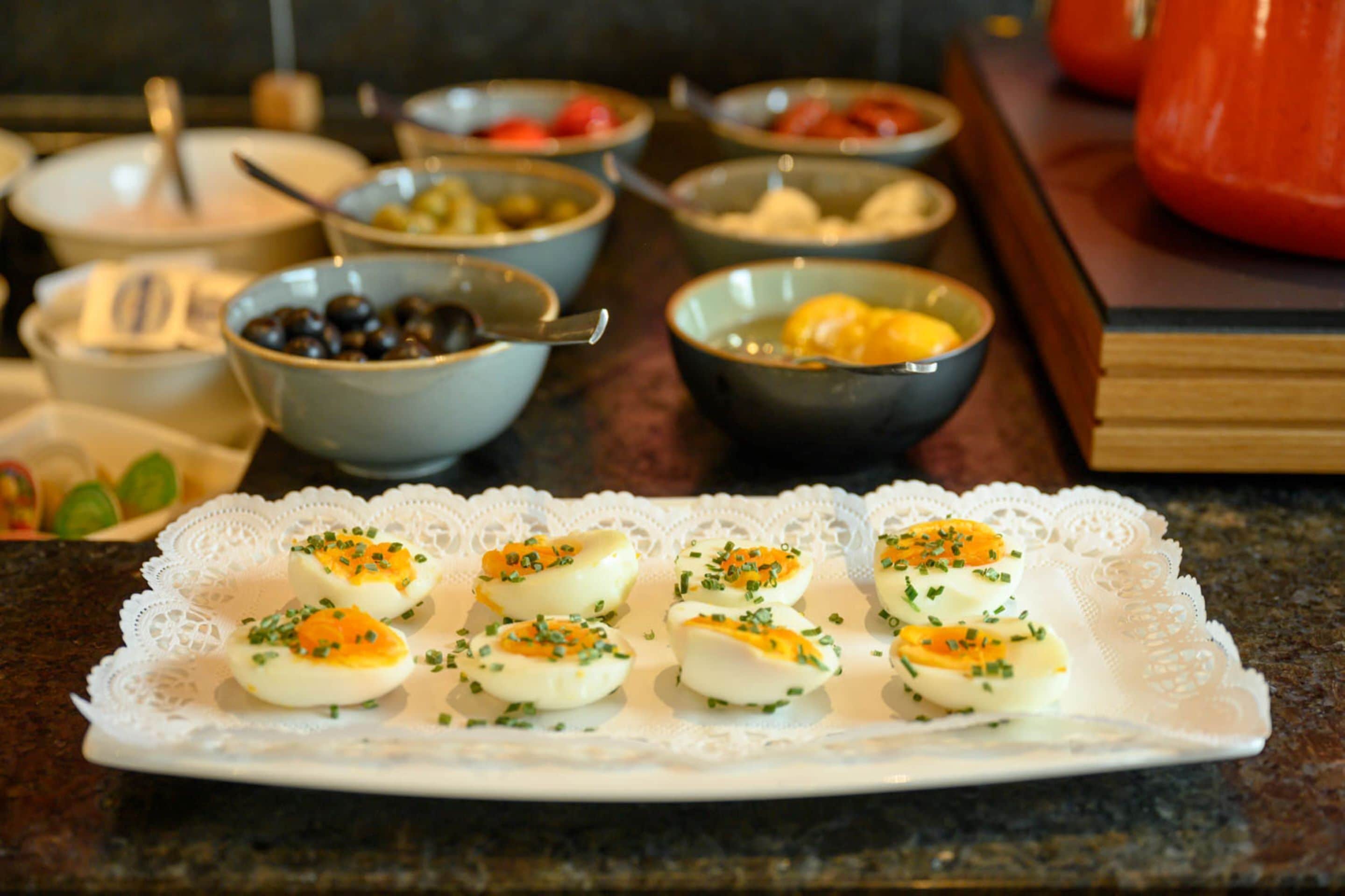 Un plato de huevos rellenos sobre un encaje de encaje, rodeado de varios recipientes con comida en una encimera de cocina.