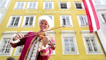 Une personne vêtue d’une tenue rouge et or joue du violon devant un bâtiment jaune sur lequel on peut lire : « Mozarts Geburtshaus ».