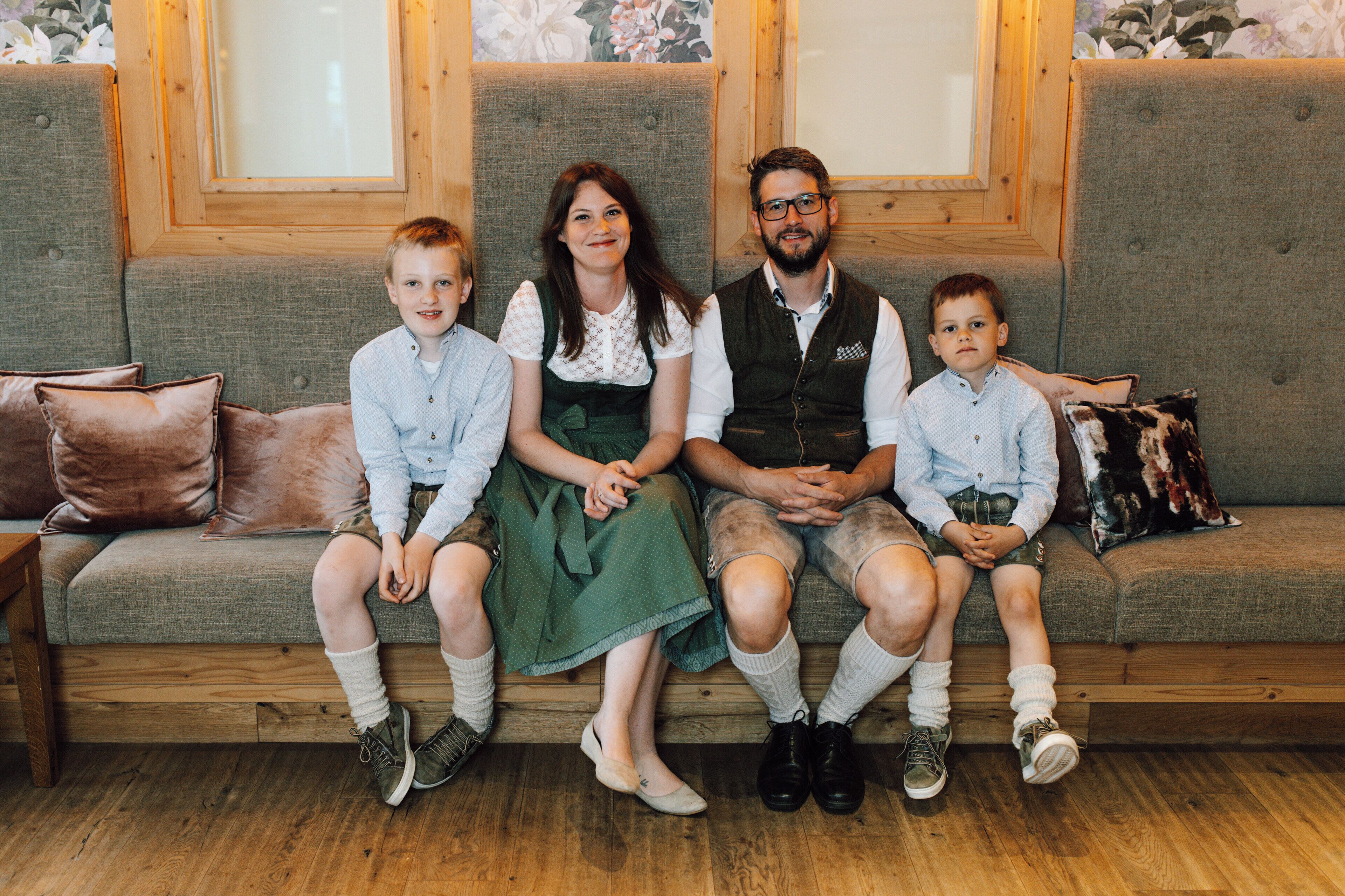 Una familia de cuatro sentada en un banco de madera con respaldo acolchado, en una habitación con paneles de madera y papel tapiz floral.