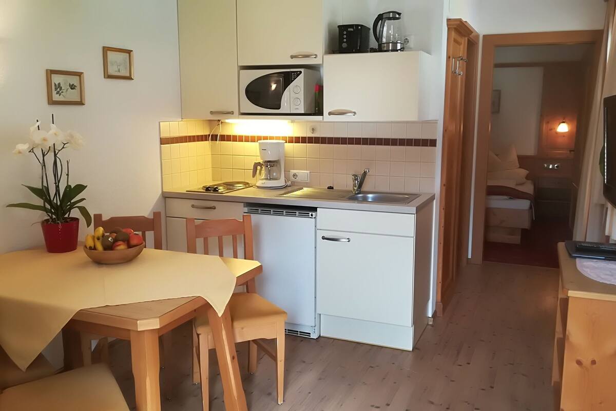Une cuisine compacte équipée d’un four à micro-ondes, d’une machine à café et d’un évier. On y trouve une table à manger avec des chaises et une plante en pot.