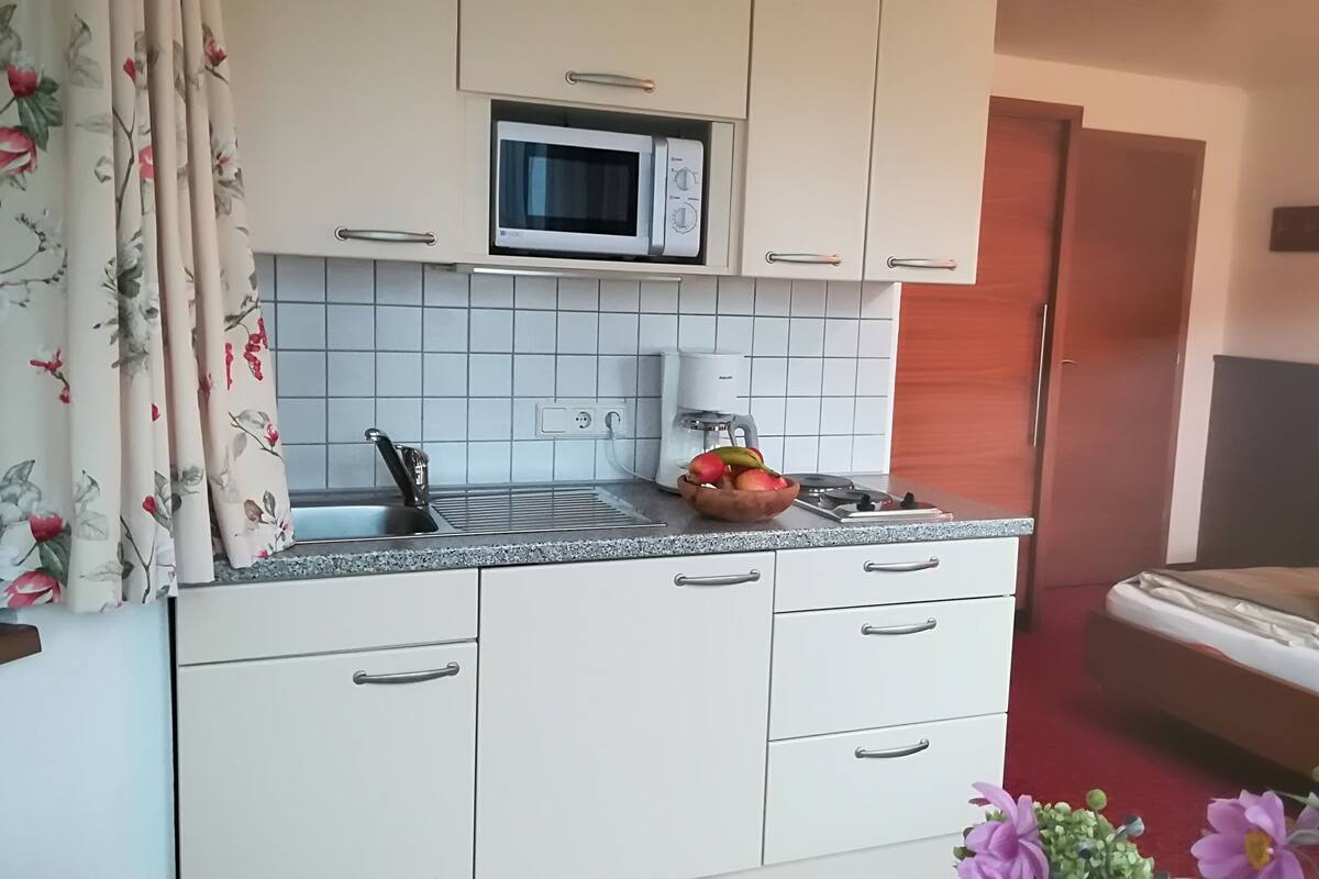 Une petite cuisine équipée d’un four à micro-ondes, d’un évier et d’une machine à café. On y trouve un petit plan de travail avec un panier de fruits et un bloc à couteaux.