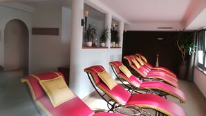 A row of sun loungers with red and gold cushions in a room with white walls, a dark ceiling, and potted plants.
