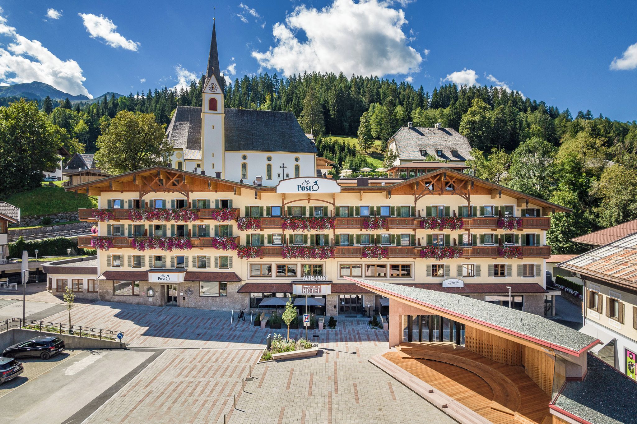 Ein Hotel mit mehreren Balkonen, die mit Blumen geschmückt sind, ein markanter Kirchturm und eine gebirgige Landschaft im Hintergrund.