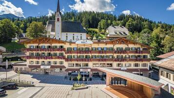 Ein Hotel mit mehreren Balkonen, die mit Blumen geschmückt sind, ein markanter Kirchturm und eine gebirgige Landschaft im Hintergrund.