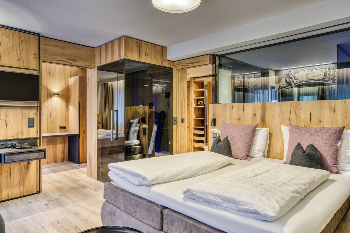 A modern bedroom with a wooden headboard, a large bed with white linens, a built-in desk, and a glass partition.