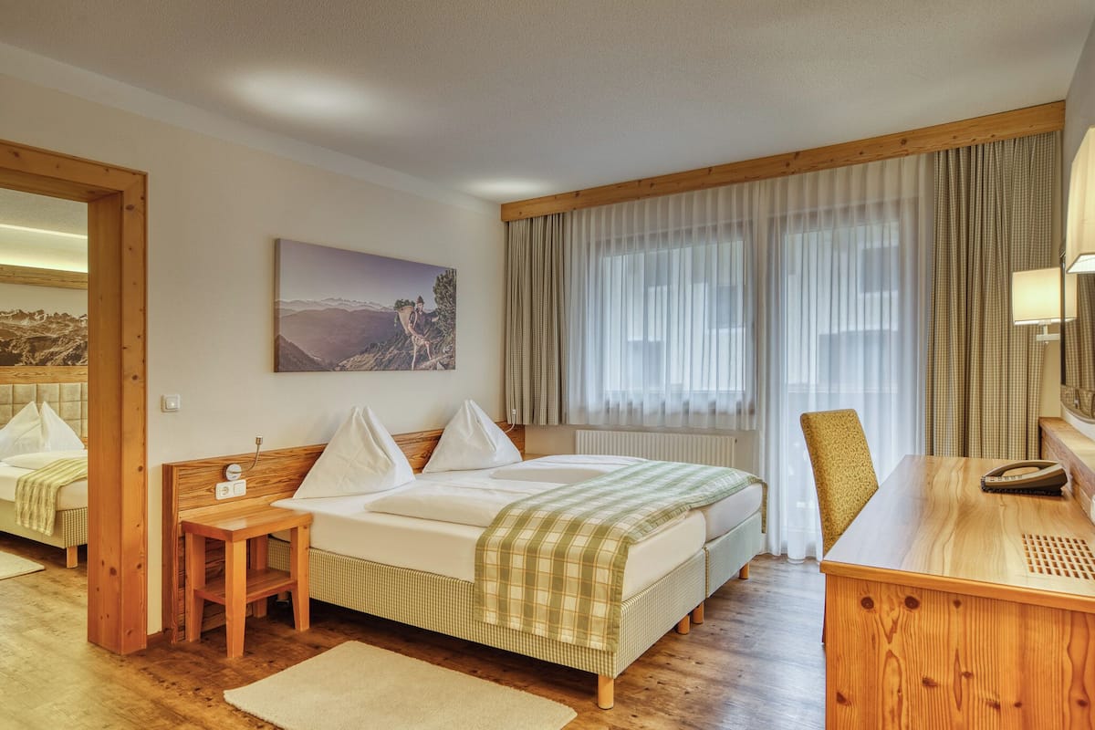 A hotel room with a bed, a wooden desk, a chair, a bedside table, a mirror, and a window with curtains.