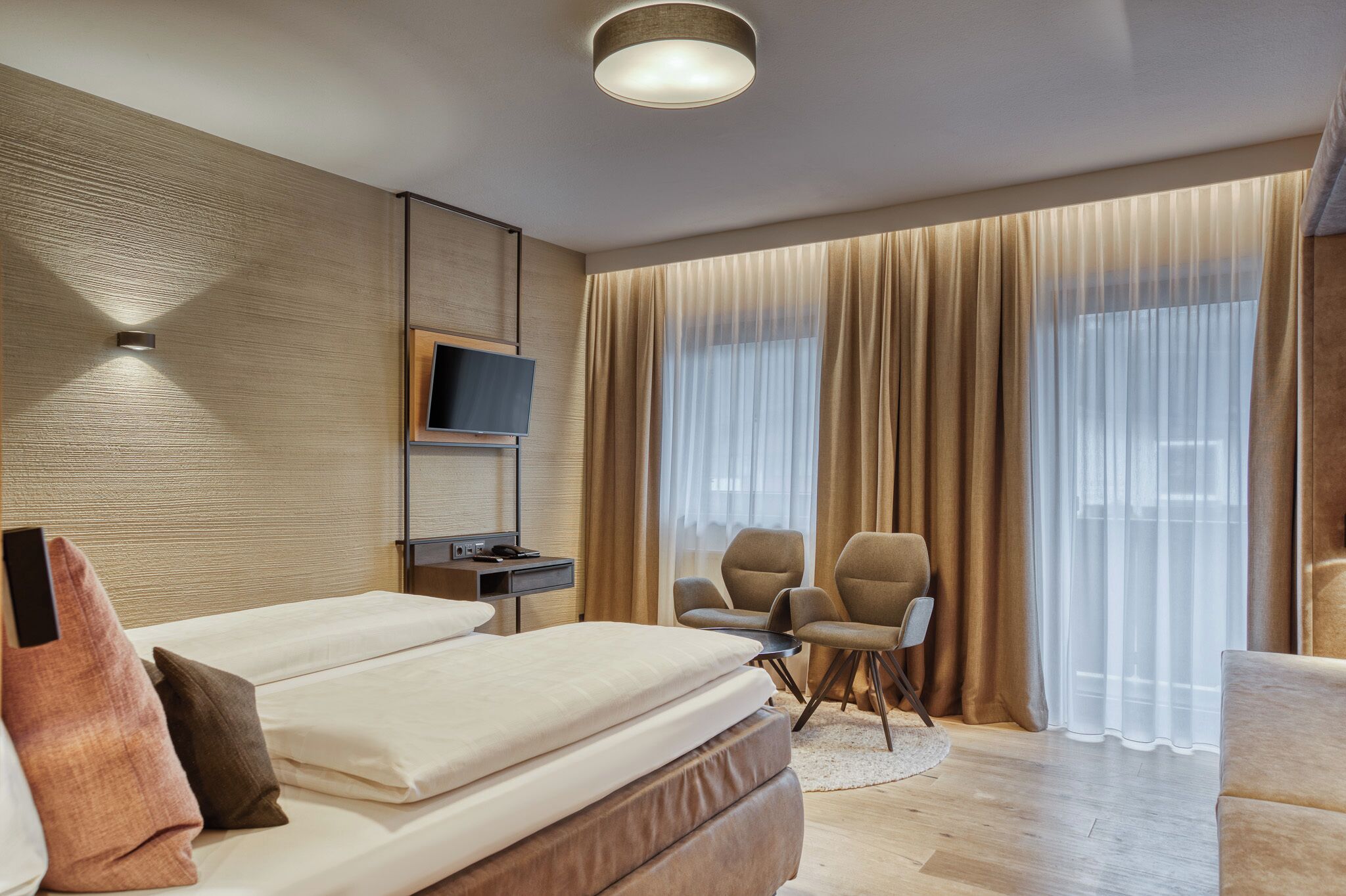 A modern hotel room with a large bed, a desk with a chair, a television, and a large window with curtains.