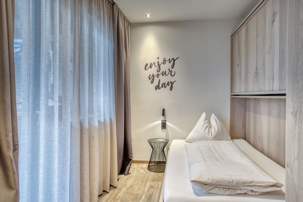 A bedroom with a bed, a wall-mounted light, a small side table, and a motivational quote on the wall.