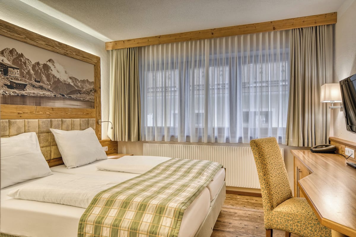 A hotel room with a bed, a chair, a desk, a lamp, and a painting on the wall.