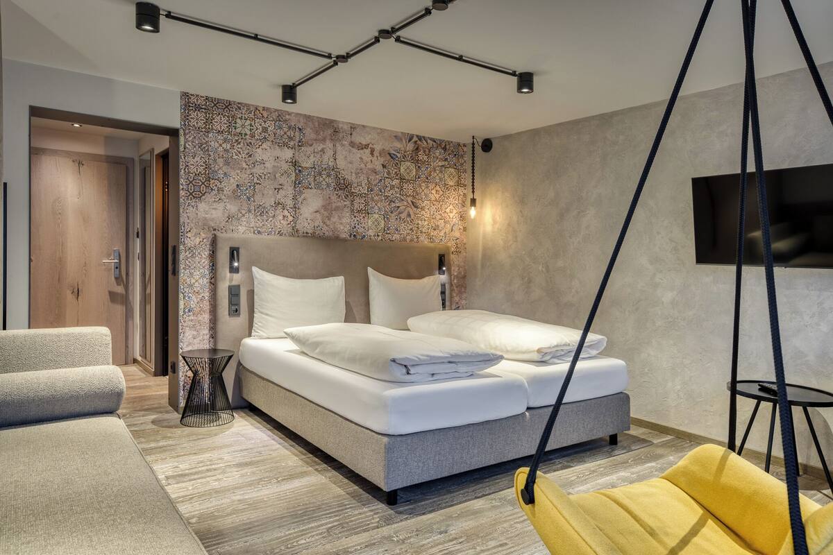 A modern bedroom with a large bed, a hanging chair, a sofa, and a TV mounted on the wall.