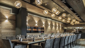 Una larga mesa de comedor preparada para una comida, con múltiples sillas, luces colgantes hexagonales en el techo y una pared de madera con estanterías empotradas al fondo.