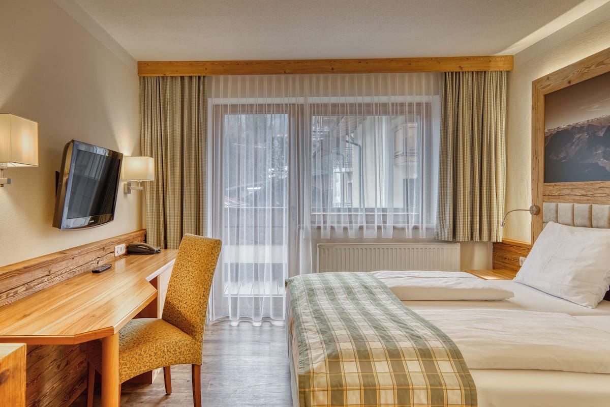 A hotel room with a wooden desk, a chair, a bed with a plaid bedspread, a television on the wall, and a window with sheer curtains.