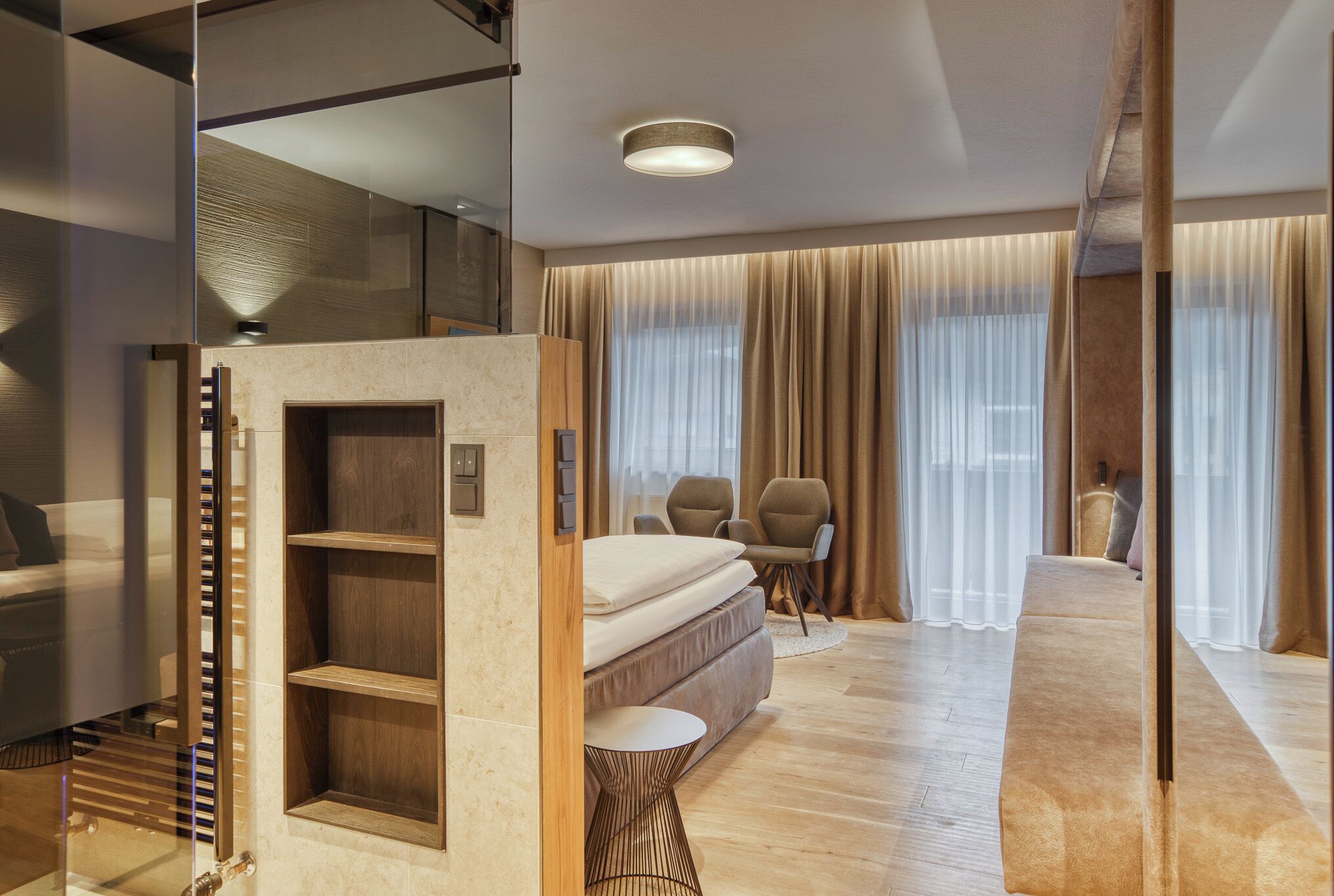 A modern hotel room with a large bed, a seating area with chairs, and a glass partition.