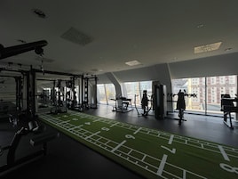 Un gimnasio con diverso equipo de ejercicio, una pista de carrera de césped artificial y grandes ventanales con vistas a edificios.