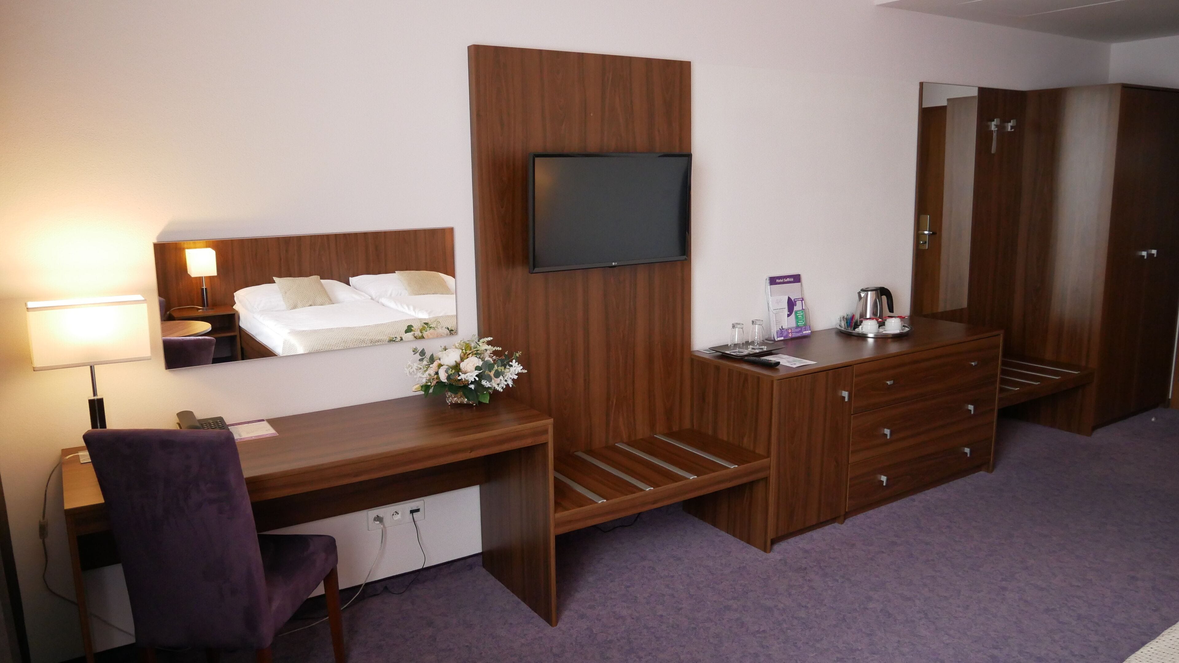 Una habitación de hotel con una cama, un escritorio con una silla, un televisor montado en la pared, un aparador y un espejo.
