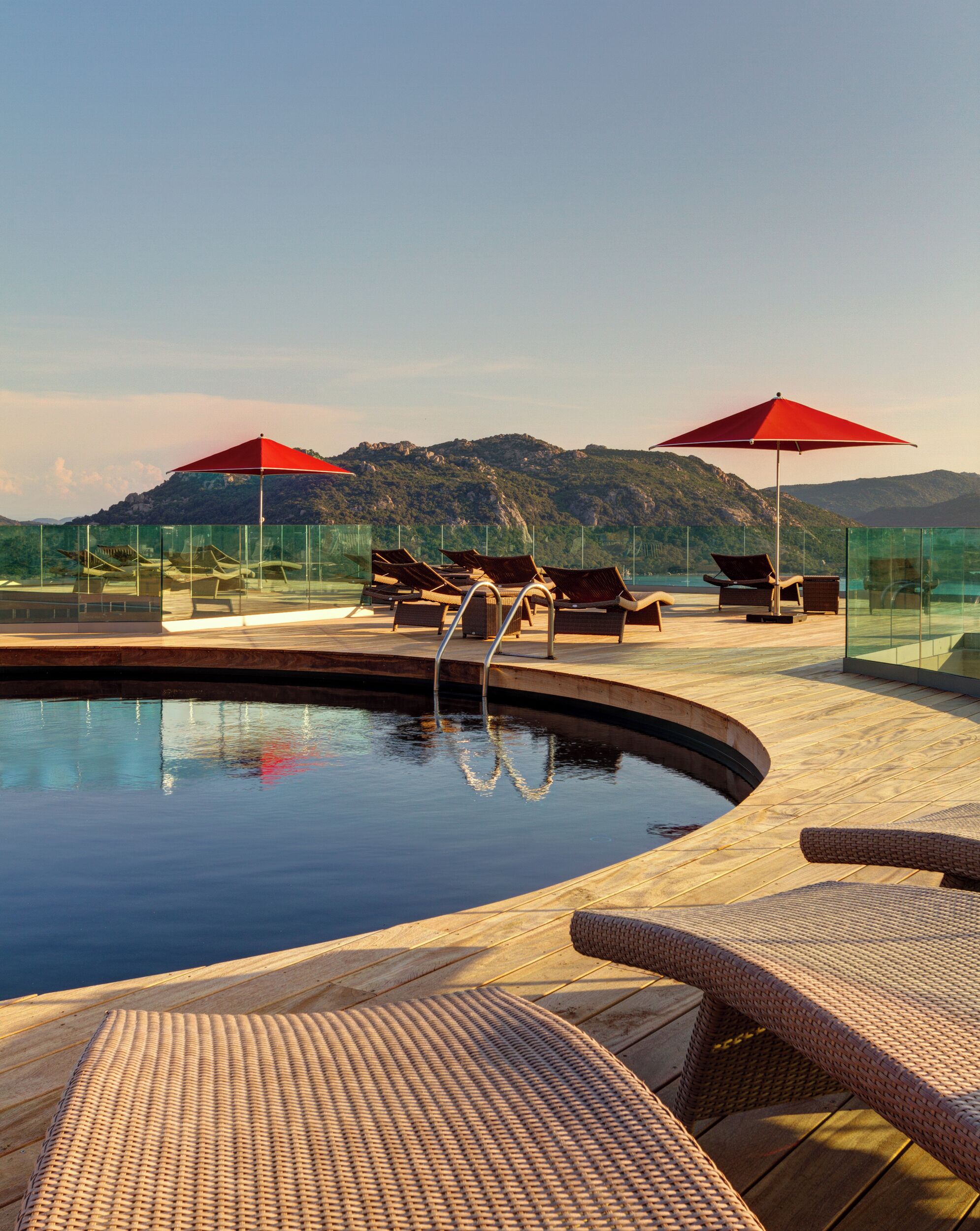A poolside area with lounge chairs and umbrellas, surrounded by glass walls and a mountainous backdrop.