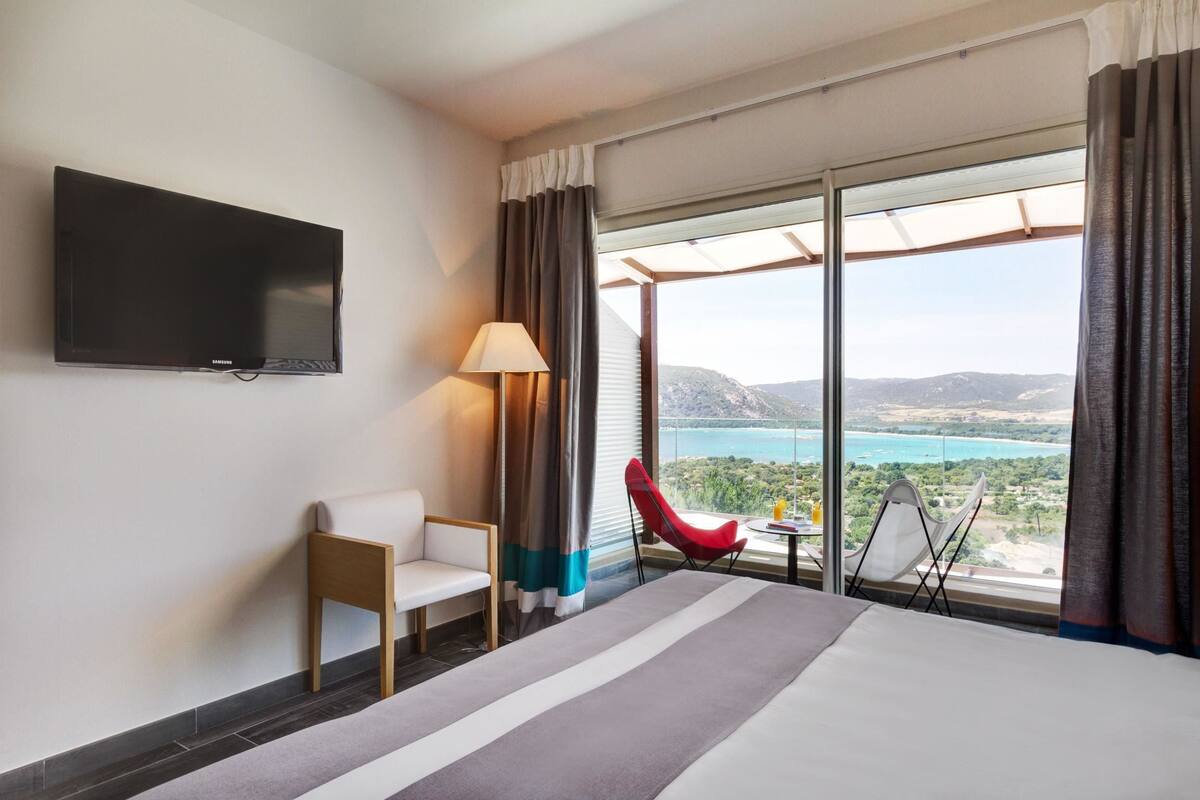 A hotel room with a bed, a TV, a chair, a table, and a view of a body of water and mountains.