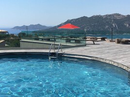 Una piscina con cubierta de madera, sillas de descanso y un paraguas rojo.