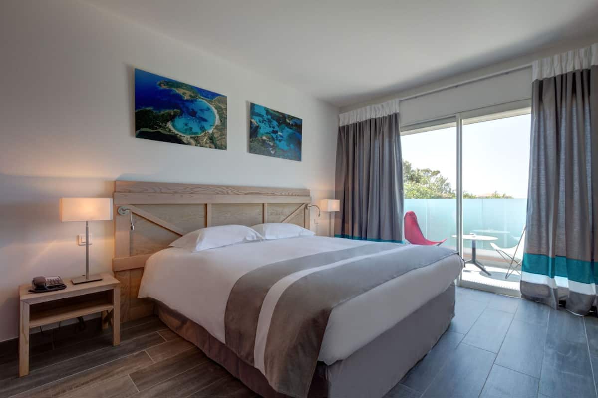 A modern hotel room with a large bed, a nightstand, a red chair, and a balcony with a view of the sea.