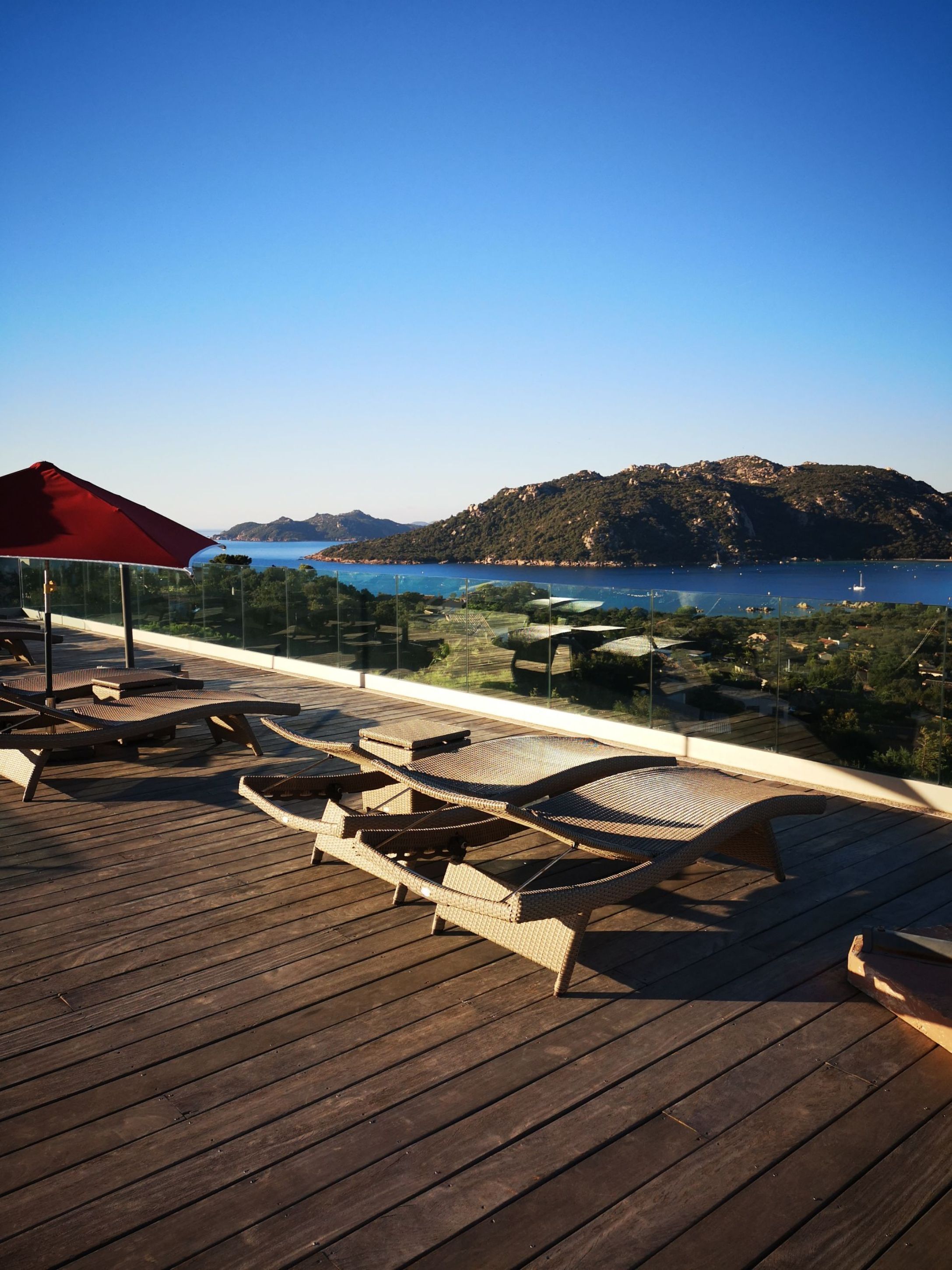 A wooden deck with lounge chairs and a red umbrella, overlooking a coastal landscape with hills and a clear blue sky.