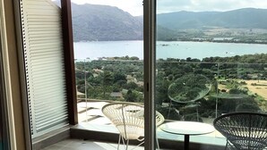 A balcony with a view of a lake and mountains, a glass table, and two chairs. - Hotel Carre Noir (Porto-Vecchio)