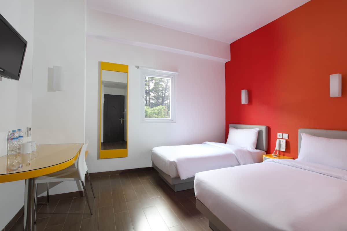Ein Hotelzimmer mit zwei Betten, einem Schreibtisch, einem Stuhl, einem Fernseher, einem Spiegel, einem Fenster und einer roten Akzentwand.