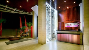 A modern hotel lobby with a reception desk, a decorative partition, and a seating area with a small table and chairs. - favehotel Kuta Square (Kuta)