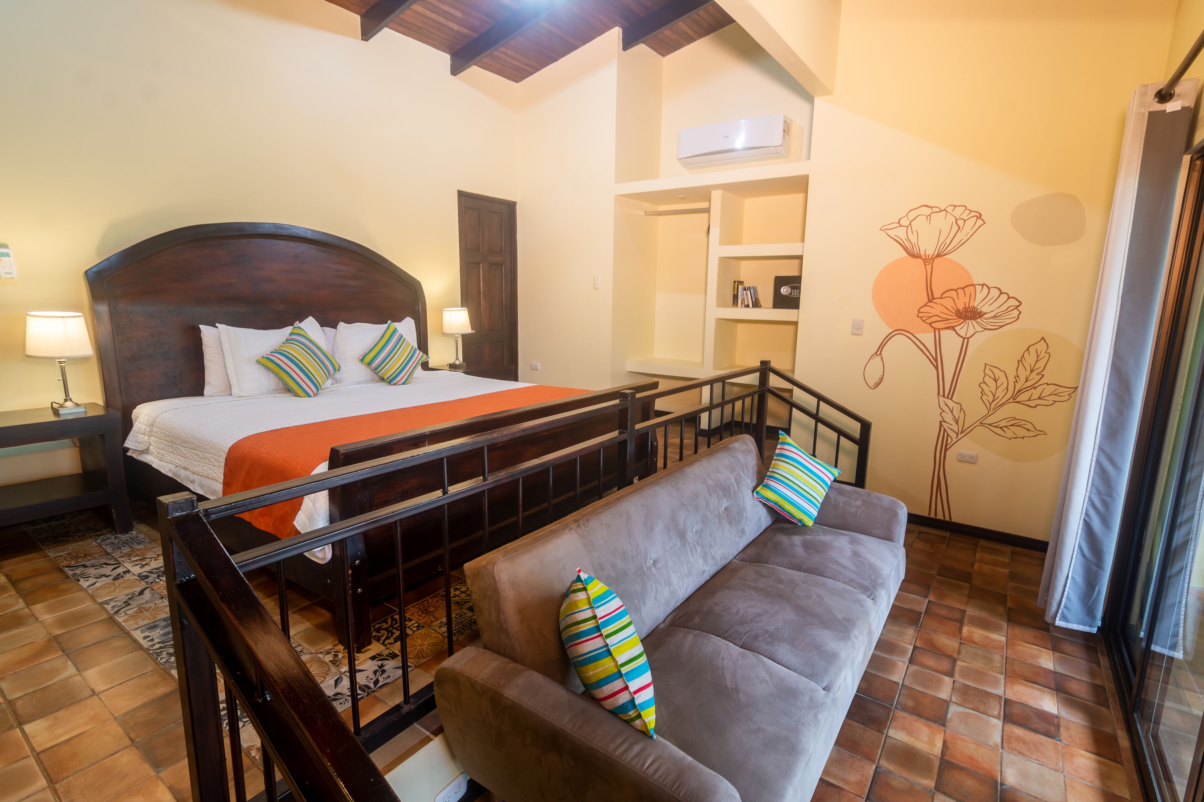 A bedroom with a bed, a sofa, a wooden headboard, a tiled floor, a wall-mounted air conditioner, and a window with curtains.