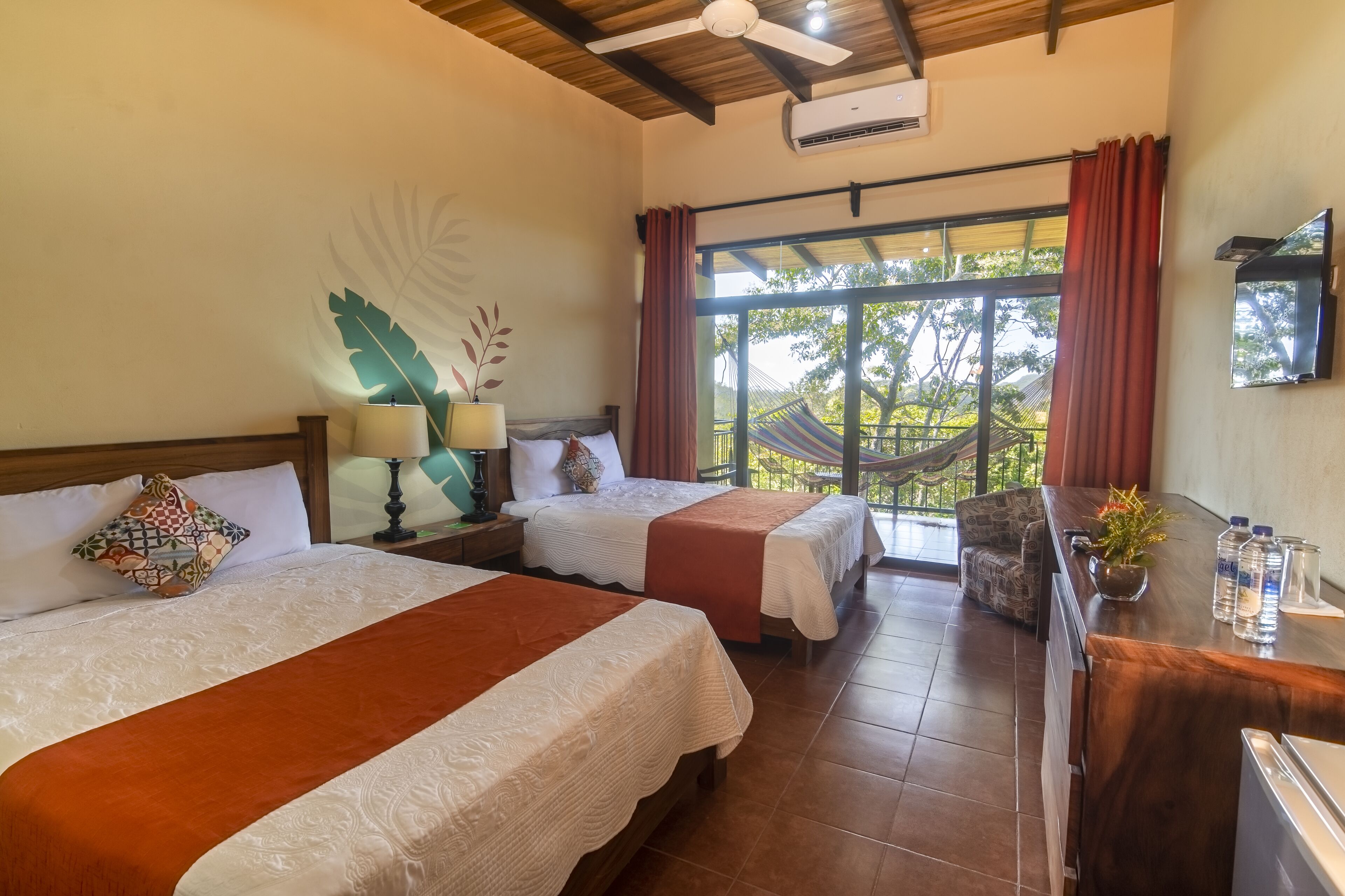 A hotel room with two beds, a desk with a mirror, a TV, and a large window with curtains.