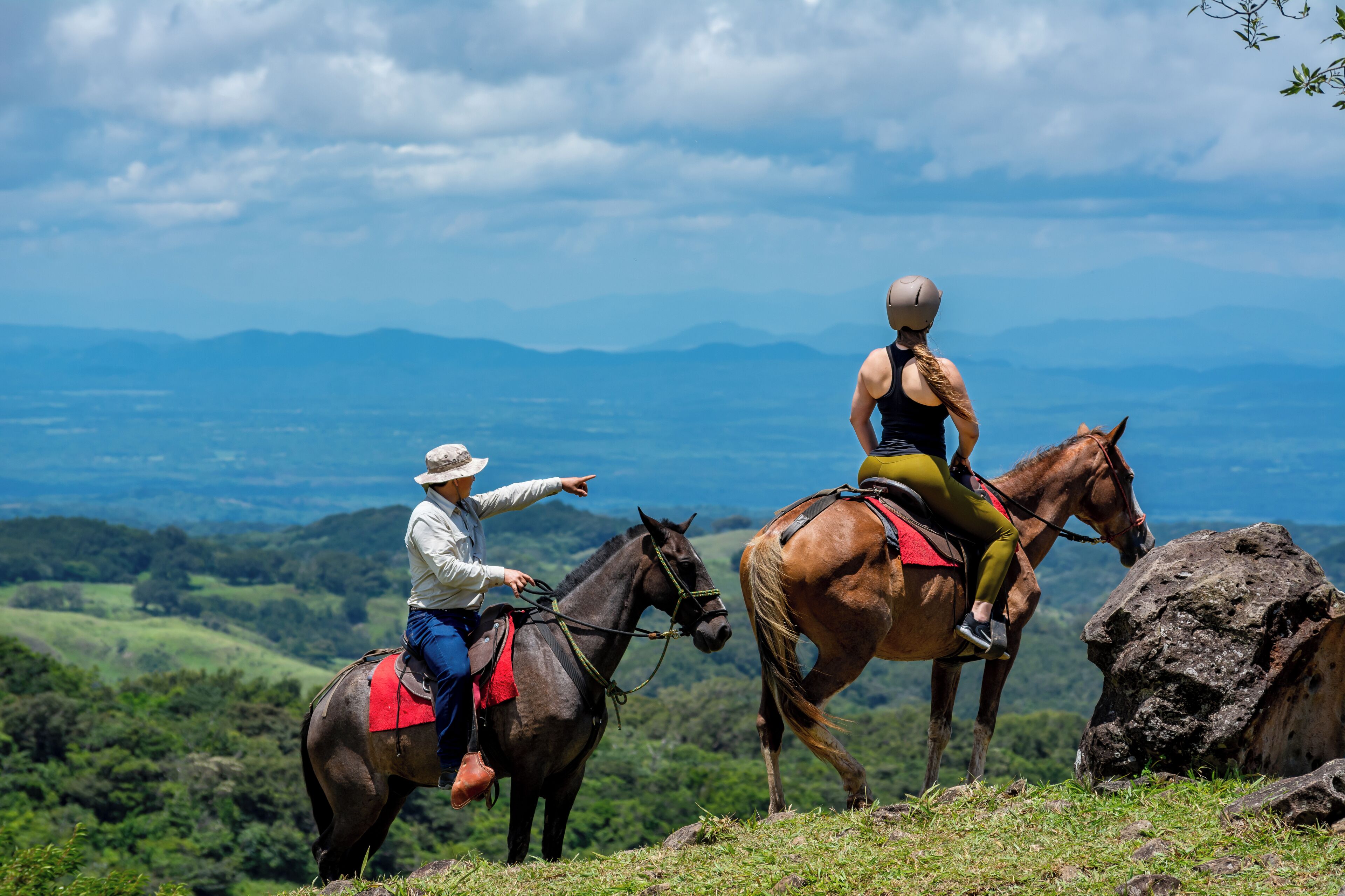 Duas pessoas cavalgam a cavalo numa colina gramada, com vista para montanhas ao longe.