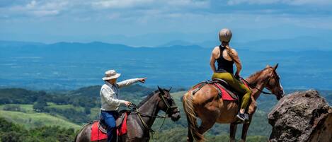 Two people riding horses on a grassy hill with a view of distant mountains.